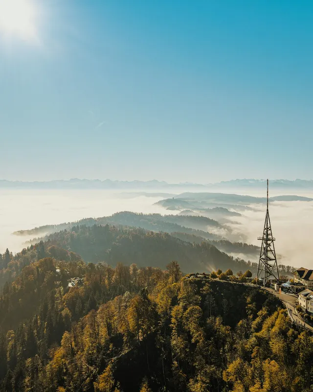 A broadcast tower and a hilltop building above a sea of fog, with distant mountains under a clear sky.