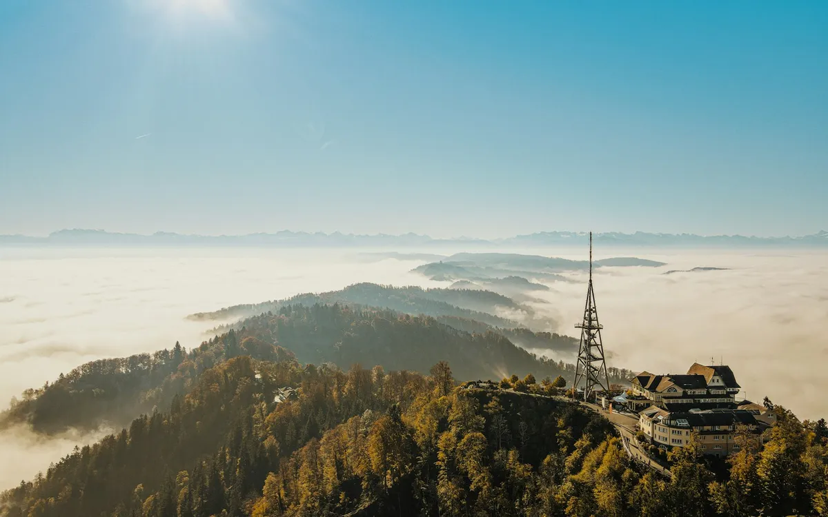 A broadcast tower and a hilltop building above a sea of fog, with distant mountains under a clear sky.