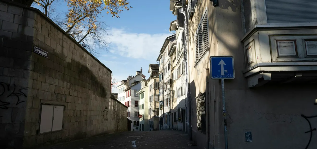 A quiet cobbled lane in Zurich between a stone wall and tall old houses, with autumn leaves on a tree overhead.