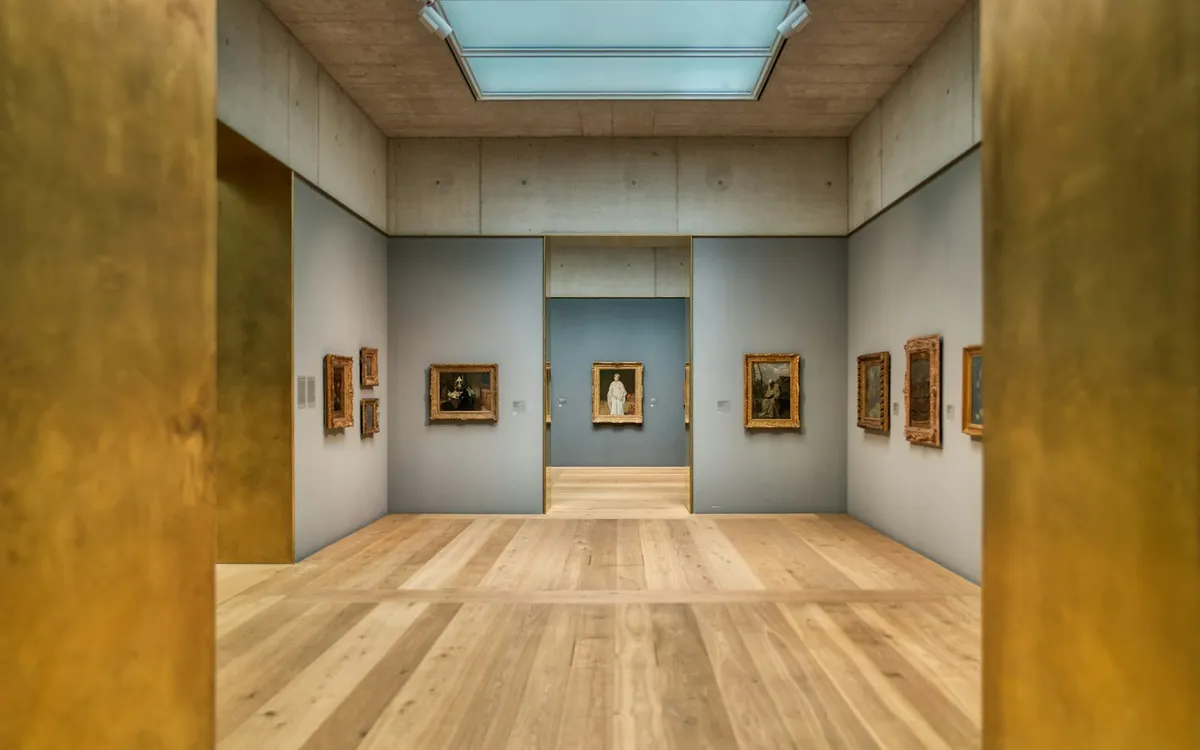 A gallery room with framed paintings on blue-grey walls, a skylight and a wooden floor.